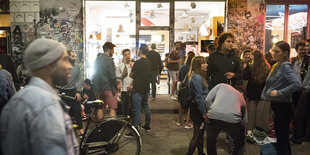 Menschen stehen in kleinen Gruppen vor einem Kiosk und trinken Bier.