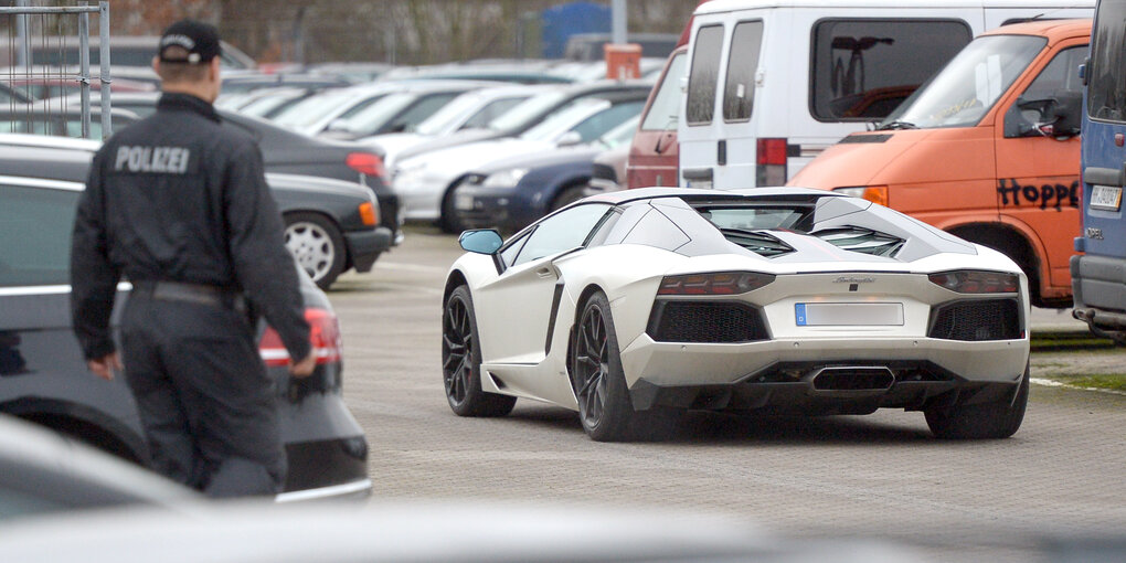 Ein weißes Sportauto steht auf einem Parkplatz, ein Polizist geht darauf zu.