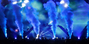 Ein DJ steht an einem erhöhten Turntable auf einer blau ausgeleuchteten Bühne, davor Bühnennebel