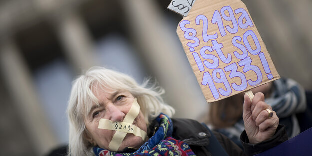 Eine Frau hat sich den Mund mit Tape verklebt, auf dem „219a“ steht. Sie hält ein Schild in der Hand mit der Aufschrift „§219a ist so 1933“