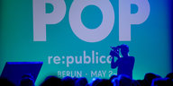 Die Silouette eines Menschen, der eine Kamera hält, vor einer türkis-blauen Wand, auf der „POP“ und „re:publica“ steht