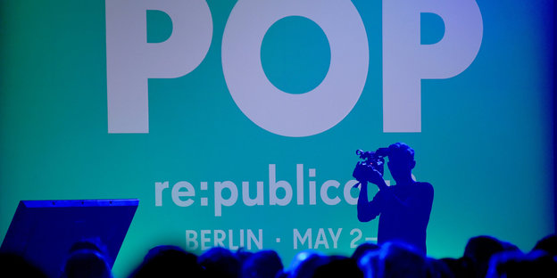 Die Silouette eines Menschen, der eine Kamera hält, vor einer türkis-blauen Wand, auf der „POP“ und „re:publica“ steht