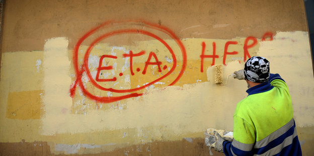 Ein Maler überpinselt eine rote Schrift auf einer gelben Hauswand. "ETA" taucht darin auf; den Rest kann man nicht lesen