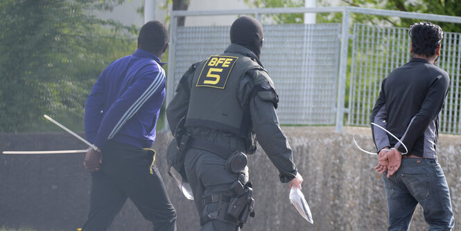 Ein maskierter Polizist läuft neben zwei mit Kabelbindern gefesselten Männern her