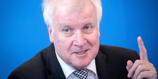 Horst Seehofer sitzt vor einem blauen Hintergrund und hebt den Zeigefinger