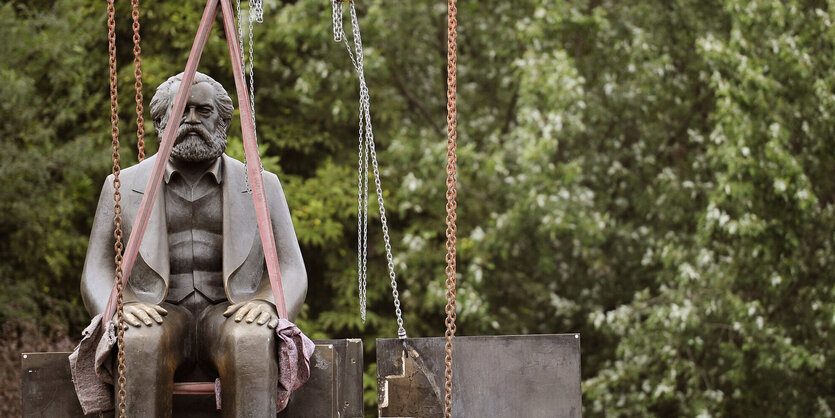 Eine Statue von einem sitzenden, älteren Mann hängt an Seilen in der Luft, im Hintergrund sieht man das Grün eines Baums.