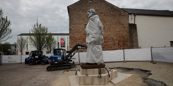 Eine in weißte Tücher eingehüllte Statue vor einer Brandwand. Der Kopf sieht durch die Verhüllung aus wie das aufgerissene Maul eines Löwen