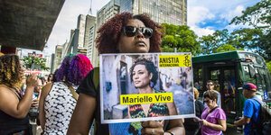 Schwarze Frau mit Sonnenbrille hält ein Schild in der Hand mit einem Foto von Marielle Franco und dem Slogan: "Marielle Vive"