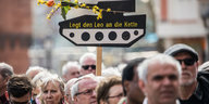 Menschen stehen nebeneinander, über ihnen ragt ein Schild in Form eines Panzers in die Höhe, auf dem „Legt den Leo an die Kette“ steht. Eine Blume ist an das Panzerrohr gebunden