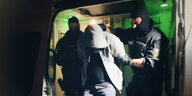 Zwei Polizisten führen einen Menschen, dessen gebeugter Kopf unter einer Kapuze verborgen ist, ab