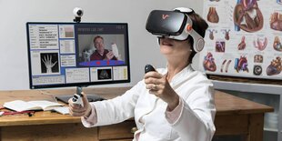 Eine Ärztin sitzt vor einem Laptop, auf dem Display ist ein Patiient zu sehen. Sie trägt eine Virtual-Reality-Brille