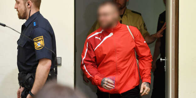 Ein Mann im roten Trainingsdress tritt hinter einem Vollzugsbeamten in den Saal, sein Gesicht ist verpixelt