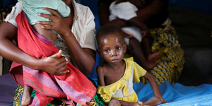 Ein unterernährtes Kind neben einer Frau, die ein weiteres Kind auf dem Arm hält.