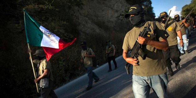 Männer mit Sturmhauben,Gewehren und einer mexikanischen Fahne laufen in Reihen durch die Straßen