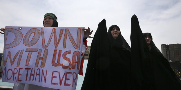Eine Frau hält ein Plakat, auf dem steht „Down with USA. More than ever“, zwei andere heben die Hand in die Höhe