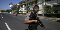 Ein Polizist im Vordergrund hält ein Gewehr und blickt zu Boden, hinter ihm ein langgestrecktes Gebäude