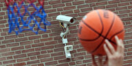 Zwei Hände, die einen Basketball halten. Im Hintergrund eine Mauer mit Überwachungskamera.