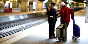 Auf einem lBahnsteig. Eine Bahnbeschäftigte unterhält sich mit einer älteren Reisenden mit Gepäck