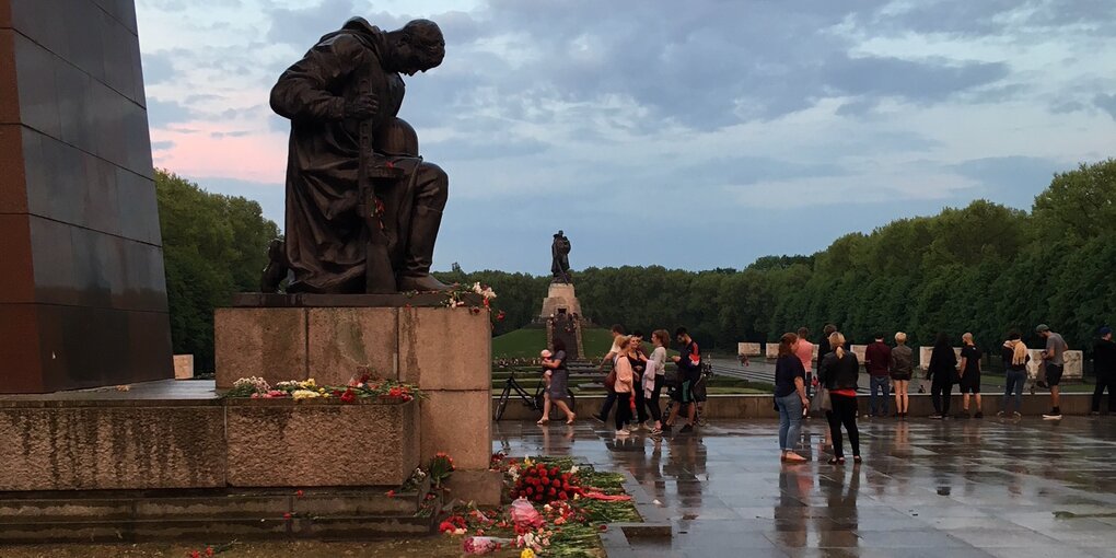 Eine Denkmalanlage, es stehen einige Menschen herum, es liegen einige Blumen herum