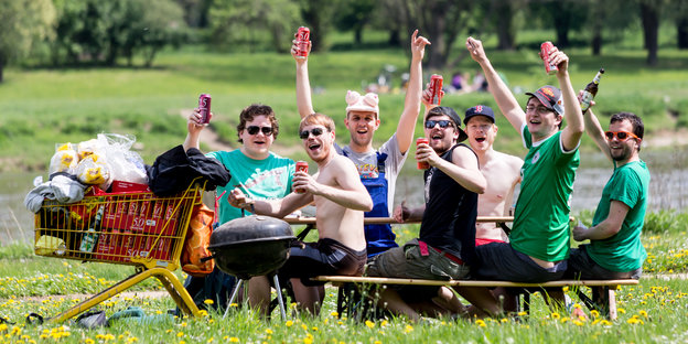 Auf Bänken sitzt eine Gruppe zum Teil unbekleideter Männer, in der Hand Bierdosen, daneben ein voller Einkaufswagen mit Bier