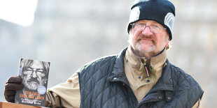 Richard Brox hat einen Arm auf die Lehne einer Bank gelegt und hält sein Buch in der Hand