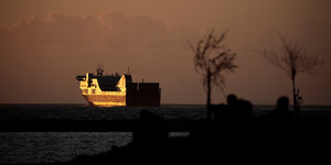 ein Schiff im Sonnenuntergang, im Vordergrund die Silhouetten von mehreren Menschen