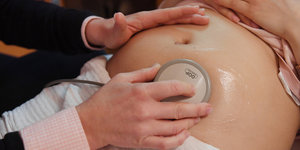 Zwei Hände führen ein Gerät zum Abhören von Herztönen über den Bauch einer Schwangeren.
