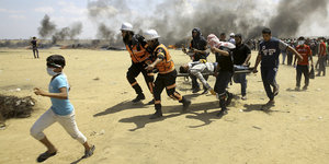 Sanitäter und andere Helfer tragen einen Verletzten auf einer Bahre über sandigen Grund, ein Junge läuft ihnen voraus. Im Hintergrund schwarzer Rauch