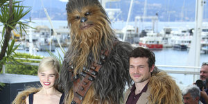 Der Schauspieler Alden Ehrenreich und die Schauspielerin Emilia Clarke stehen neben einer Person in einem Kostüm von Chewbacca