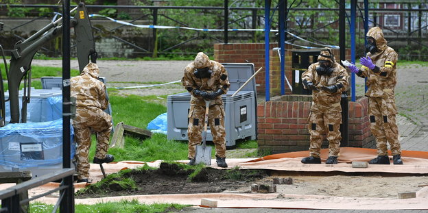 Vier Männer in Militärkleidung und Schutzmasken untersuchen den Boden. Im Hintergrund große graue Boxen.