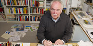 Der Karikaturist Dieter Hanitzsch sitzt mit einem Stift in der Hand vor einer Bücherwand an seinem Schreibtisch