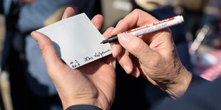 Eine Hand hält ein weißes Stück Papier, eine andere schreibt mit Edding darauf