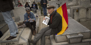 Ein Mann sitzt auf einem Grabstein und trägt die Fahne der spanischen 2. Republik über der Schulter, in der Hand hält er eine schwarz-weiß Fotografie