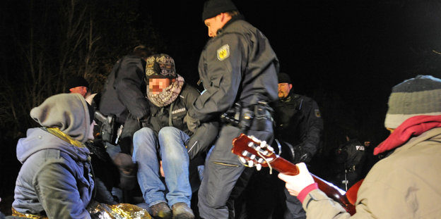 Zwei Polizisten tragen einen Aktivisten weg, zwei Aktivisten sitzen daneben, einer davon spielt Guitarre