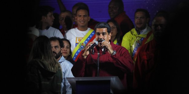 Nicolas Maduro steht hinter einem Stehpult und gestikuliert, um ihn herum Unterstützer. Von allen Seiten verdunkelt sich das Bild
