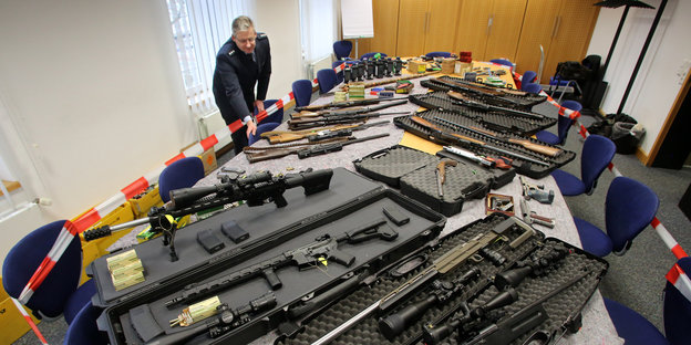 Auf einem langen Tisch liegen verschiedene Arten von Waffen. Im Hintergrund ein Mann in Polizeikleidung.