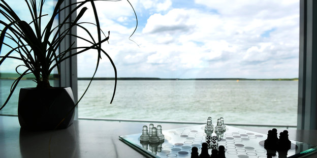 An einem Fenster mit Seeblick steht eine Topfpflanze und gläsernes Schachbrett mit Figuren