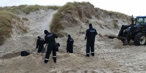 Sechs Polizisten mit Schaufeln und Bagger graben in einer Düne