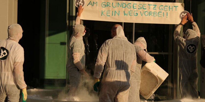 Menschen in weißen Schutzanzügen vor einer Eingangstür. Zwei von ihnen befestigen ein Transparent mit der Aufschrift „Am Grundgesetz geht kein Weg vorbei“. Auf dem Boden dampfen Eisblöcke