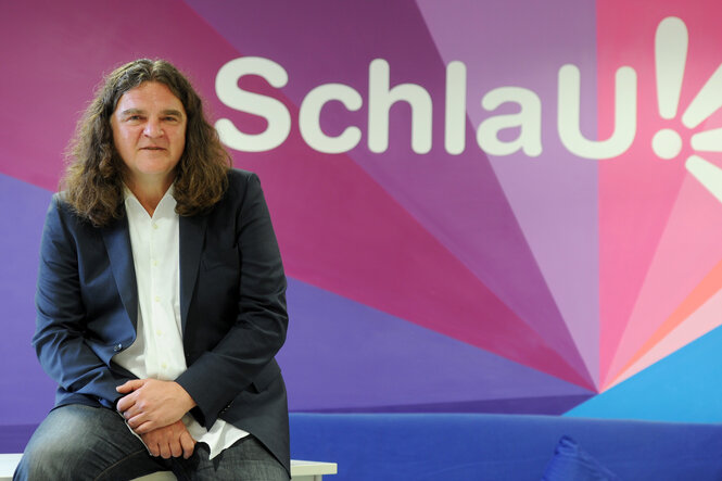 Ein Mann sitzt vor einer bunten Wand mit der Aufschrift „Schlau!“