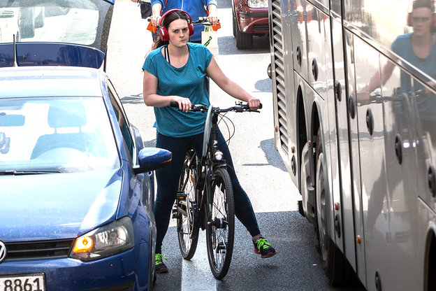 Eine Fahrradfahrerin steht eingequetscht zwischen einem Auto und einem Bus und blickt genervt