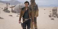 Ein Mann, es ist Han Solo, und ein Zweibeiner mit viel Fell laufen über einen windigen Wüstenplaneten