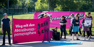 Demonstrantinnen stehen mit Plakaten vor einem Zaun