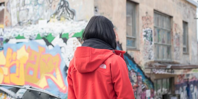 Eine Frau steht in einer roten Jacke mit dem Rücken zur Kamera vor einem Haus