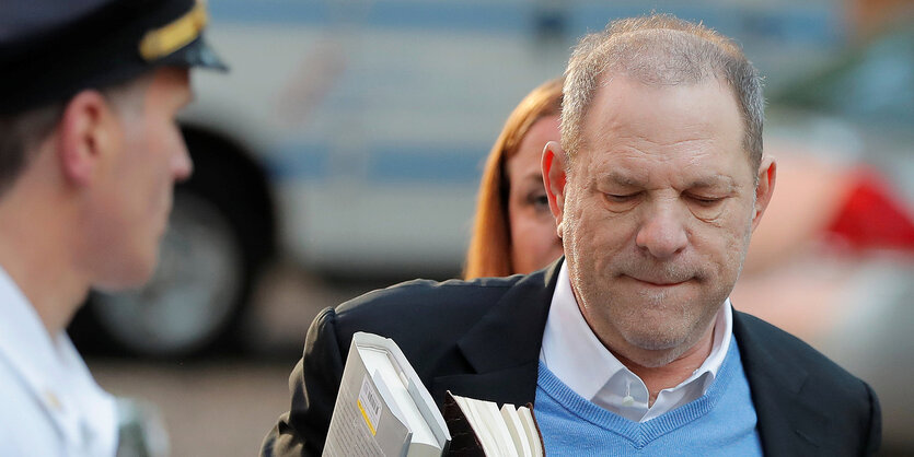 Harvey Weinstein neben einem Polizisten. Er trägt Bücher in der rechten Hand