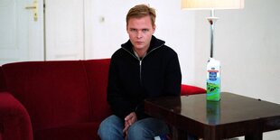 Ein jungen Mann sitzt auf einem roten Sofa, auf dem Tisch neben ihm steht ein Milchkarton