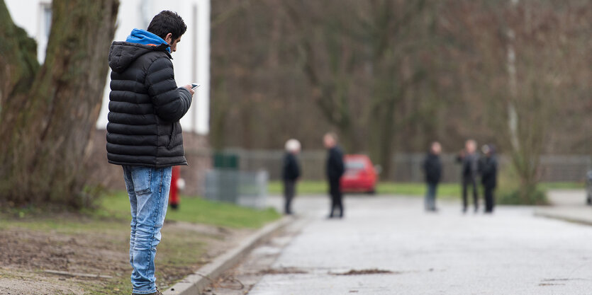 Ein Mann steht mit Smartphone am Wegesrand, weiter hinten sind unscharf weitere Personen zu sehen.