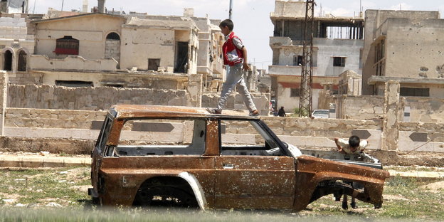 Kinder spielen auf einem Autofrack, im Hintergrund sind zerstörte Häuser zu erkennen.