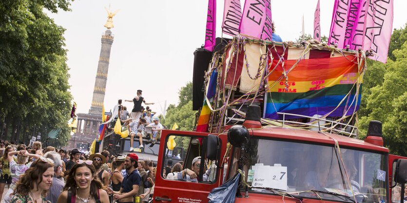 Viele Menschen, ein großes Auto mit Regenbogen- und pinken Fahnen, im Hintergrund die Siegessäule