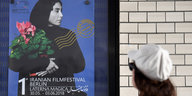 Man sieht das Plakat des 1. Iranischen Filmfestivals, darauf ist eine Frau in schwarz zu sehen, die einen Blumenstrauß trägt, der Hintergrund ist blau. Darunter stehen die Daten (30. Mai bis 3. Jun). EIne Frau schaut auf das Plakat